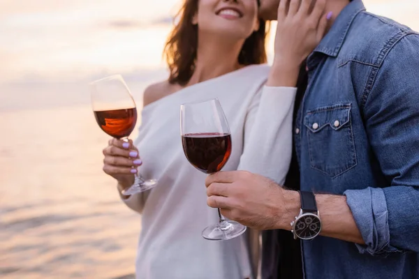 Cropped view of woman touching boyfriend while holding glass of wine near sea at sunset — Stock Photo