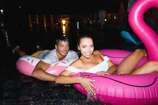 Selective focus of young couple looking at camera while swimming on rings in pool at night — Stock Photo