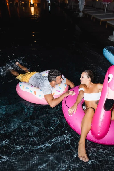 Vue grand angle de l'homme regardant petite amie sur anneau de natation dans la piscine la nuit — Photo de stock
