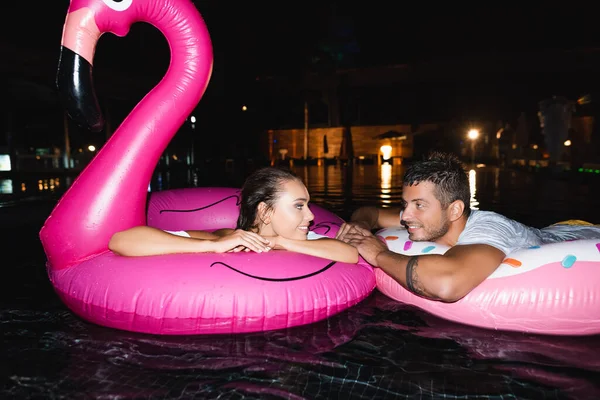 Focus selettivo di giovani coppie che si guardano l'un l'altro sugli anelli di nuoto in piscina di notte — Foto stock