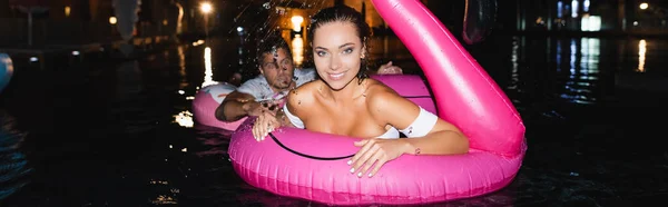 Encabezado del sitio web de la mujer joven mirando a la cámara mientras nada con anillo inflable cerca de novio en la piscina por la noche - foto de stock