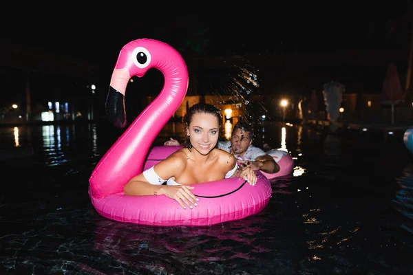 Concentration sélective de la jeune femme regardant la caméra tout en nageant dans l'anneau près du petit ami dans la piscine la nuit — Photo de stock