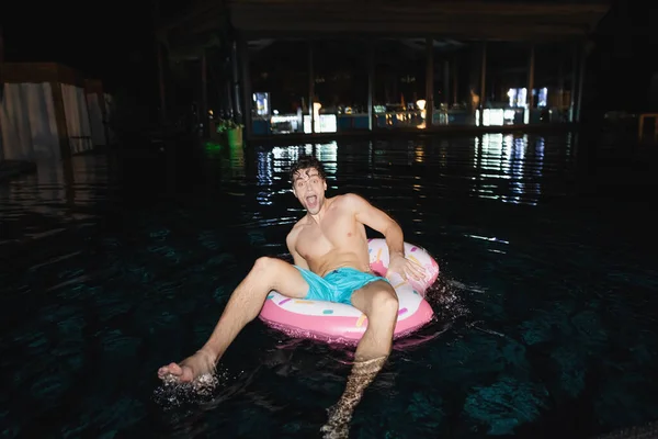 Enfoque selectivo del hombre emocionado sentado en el anillo de natación en la piscina por la noche - foto de stock