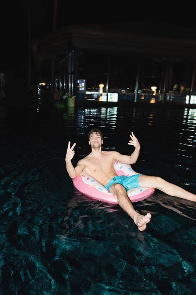 Enfoque selectivo del hombre sin camisa mostrando signo de paz en el anillo de natación en la piscina por la noche - foto de stock