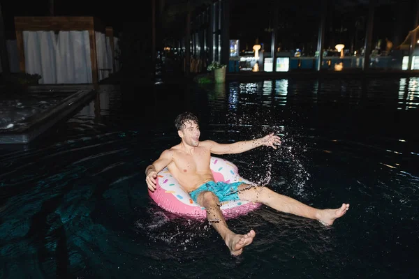 Selective focus of shirtless man on swim ring splashing water in pool at night — Stock Photo