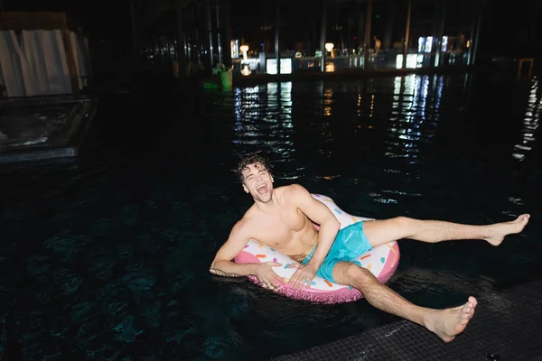Excited muscular man sitting on swim ring in pool at night — Stock Photo