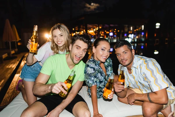 Foco seletivo de amigos com garrafas de cerveja olhando para a câmera ao ar livre à noite — Fotografia de Stock