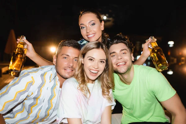 Young woman looking at camera while holding bottles of beer near friends at night — Stock Photo