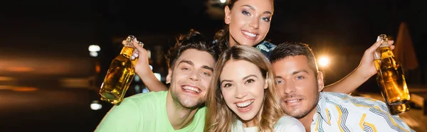 Orientação panorâmica de jovens amigos com garrafas de cerveja olhando para a câmera ao ar livre à noite — Fotografia de Stock