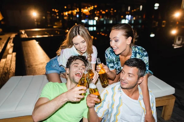 Jovens amigos brindam com garrafas de cerveja perto da piscina à noite — Fotografia de Stock