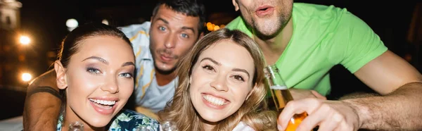 Panoramic crop of women looking at camera near boyfriends with beer at night — Stock Photo