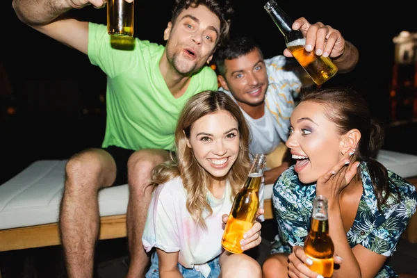 Foco seletivo de jovens amigos animados segurando garrafas de cerveja durante a festa à noite — Fotografia de Stock