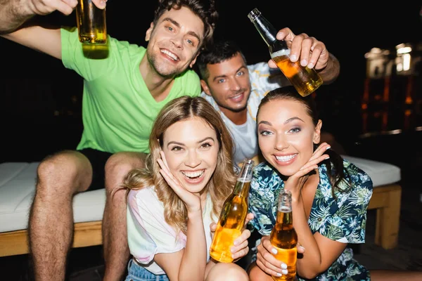 Enfoque selectivo de las mujeres con botellas de cerveza mirando a la cámara cerca de los novios por la noche - foto de stock