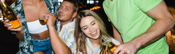 Panorâmica tiro de mulher clinking cerveja com namorado perto de amigos ao ar livre à noite — Fotografia de Stock