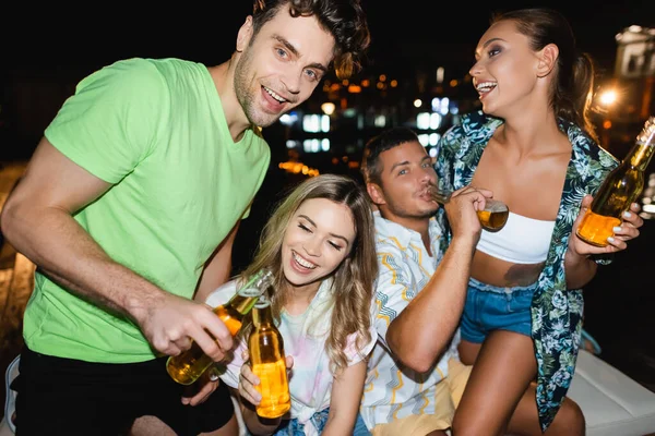 Selective focus of couple toasting with beer bottles during party with friends outdoors at night — Stock Photo