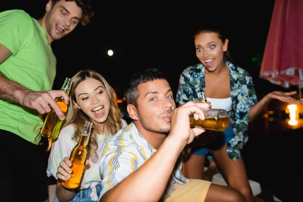 Selective focus of young man drinking beer near excited friends at night — Stock Photo