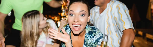 Panoramic shot of young woman looking at camera while friends toasting with beer at night — Stock Photo