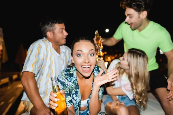 Foco seletivo da mulher animada com garrafa de cerveja olhando para a câmera perto de amigos ao ar livre à noite — Fotografia de Stock