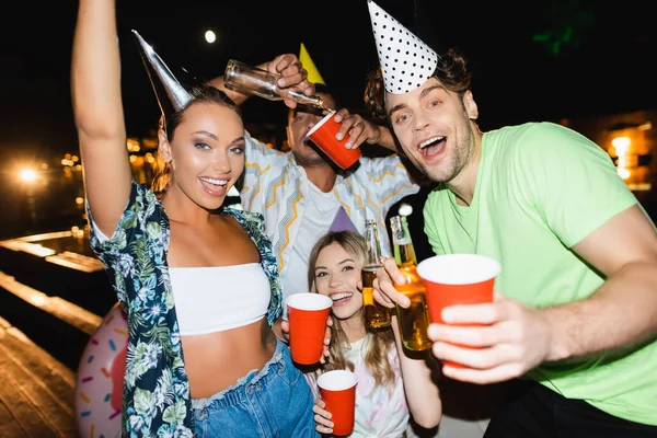 Foco seletivo de jovens em bonés de festa segurando garrafas de cerveja e copos descartáveis ao ar livre à noite — Fotografia de Stock