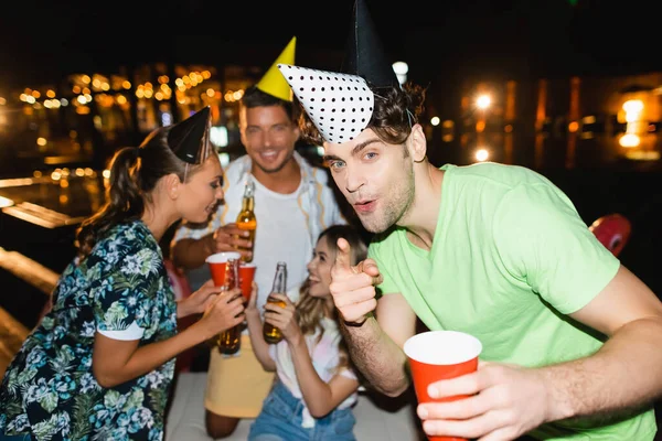 Selective focus of young man in party cap pointing with finger at camera near friends with beer outdoors at night — Stock Photo