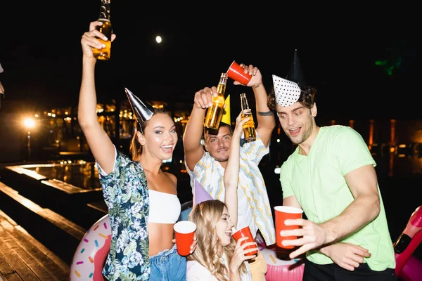 Young friends in party caps holding bottles of beer outdoors at night — Stock Photo