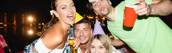 Panoramic shot of young people in party caps looking at camera during party with beer at night — Stock Photo