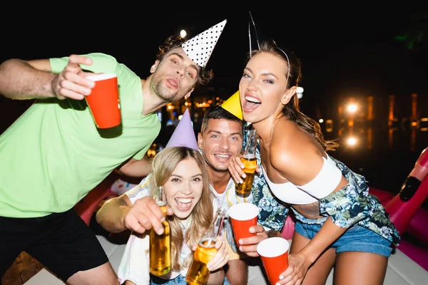 Foco seletivo de jovens amigos em bonés de festa segurando garrafas de cerveja e copos descartáveis à noite — Fotografia de Stock