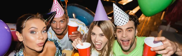 Panoramic orientation of excited friends in party caps holding disposable cups near balloons at night — Stock Photo