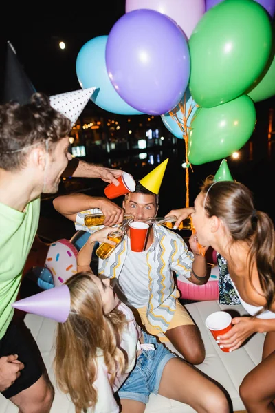 Foco seletivo do homem segurando copo descartável perto de amigos derramando cerveja e balões ao ar livre à noite — Fotografia de Stock