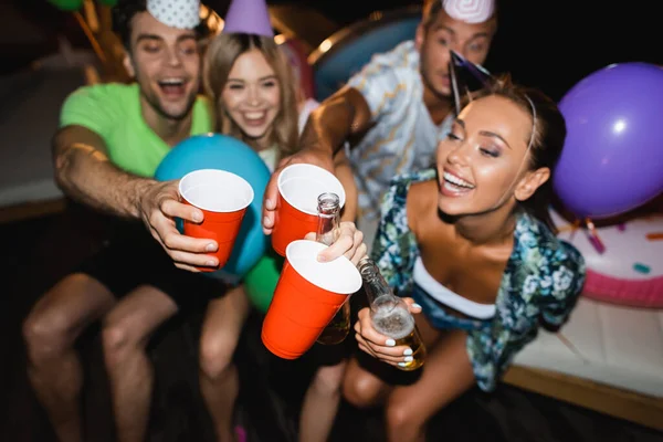 Selektiver Fokus junger Freunde in Partykappen, die nachts mit Bier und Einwegbechern klingeln — Stockfoto