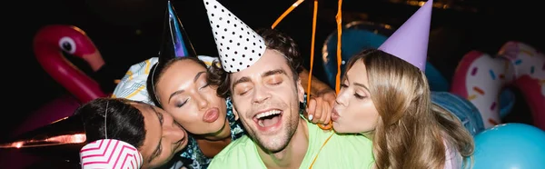 Panoramic crop of young friends kissing man in party cap near balloons at night — Stock Photo