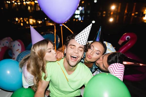 Selective focus of friends in party caps kissing man with balloons outdoors at night — Stock Photo