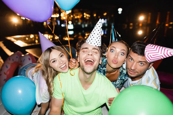 Enfoque selectivo de jóvenes amigos mirando la cámara mientras sostienen globos cerca de la piscina por la noche - foto de stock