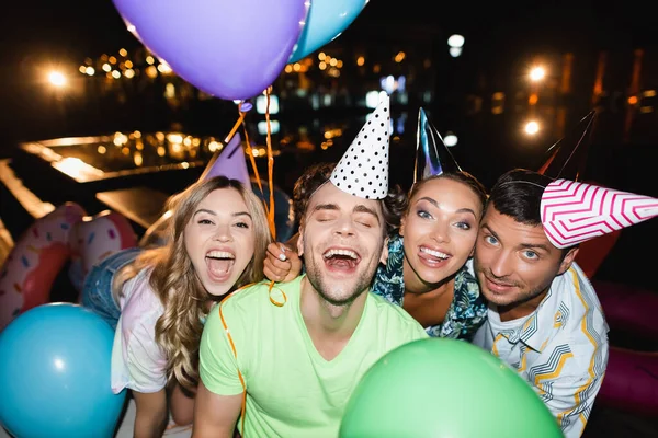 Foco seletivo de jovens em bonés de festa segurando balões à noite — Fotografia de Stock