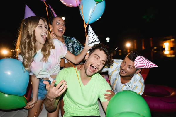 Selective focus of young people holding balloons and paper caps near friend at night — Stock Photo