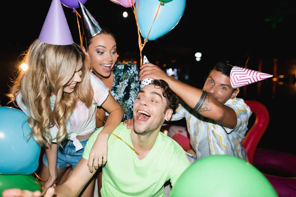 Selective focus of man wearing party cap on head of friend near women and balloons at night — Stock Photo