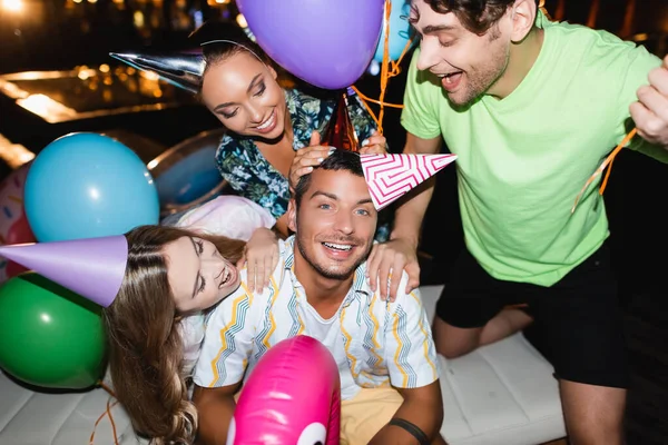 Selective focus of friends hugging young man in party cap near balloons and swimming pool at night — Stock Photo