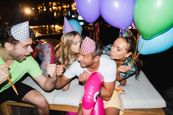 Selective focus of young men showing yeah gesture near girlfriends in party caps and balloons at night — Stock Photo