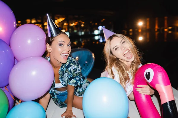 Foco seletivo de mulheres jovens em bonés de festa olhando para a câmera perto de balões à noite — Fotografia de Stock