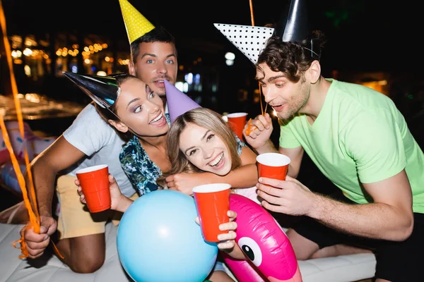 Selective focus of friends in party caps holding disposable cups near balloons at night — Stock Photo