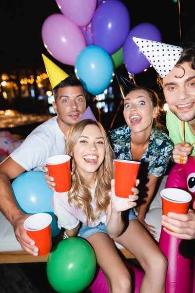 Focus selettivo di amici che tengono tazze usa e getta durante la festa con palloncini di notte — Foto stock