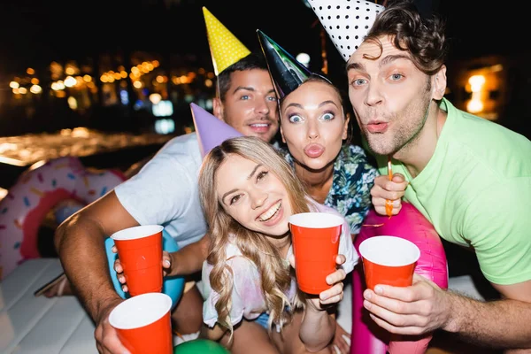 Foco seletivo de casais em bonés de festa olhando para a câmera enquanto segurando copos descartáveis à noite — Fotografia de Stock