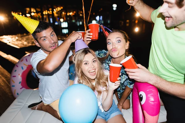 Focus selettivo di giovani amici in berretti da festa divertirsi e tenere le tazze usa e getta di notte — Foto stock