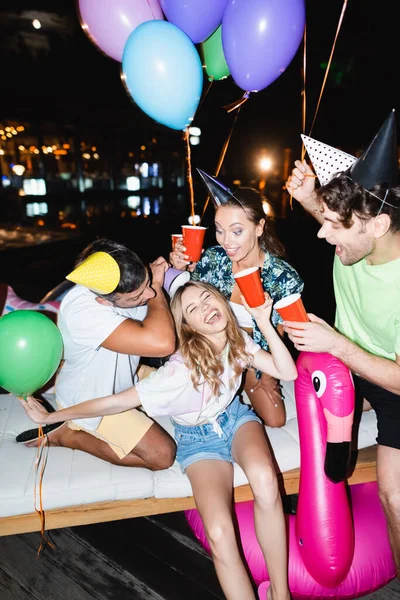 Focus selettivo della donna che tiene la tazza usa e getta vicino agli amici in berretti e palloncini di festa di notte — Foto stock