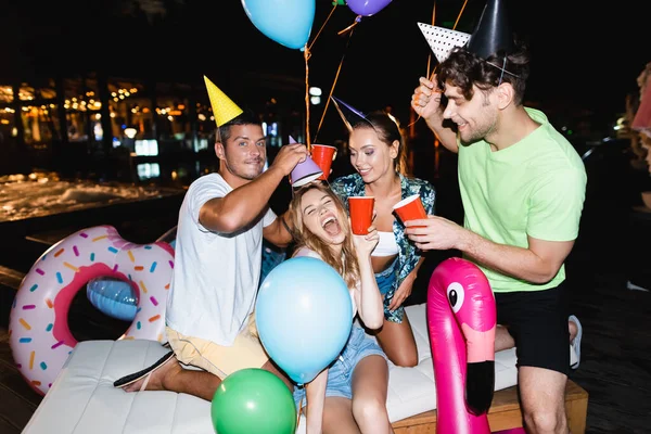 Friends in party caps holding disposable cups near swimming pool at night — Stock Photo