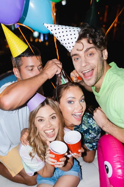 Selective focus of friends with disposable cups looking at camera near balloons outdoors at night — Stock Photo