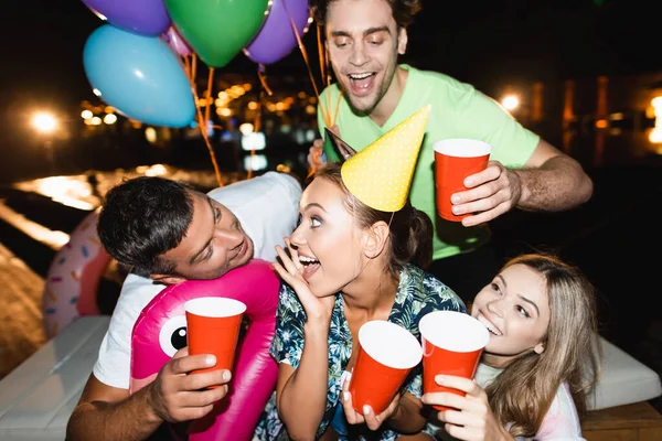 Concentration sélective de la jeune femme en bonnet de fête tenant tasse jetable près des amis avec des ballons la nuit — Photo de stock
