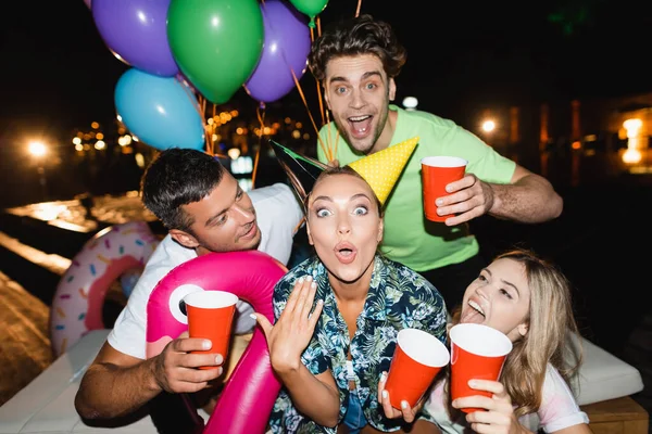 Aufgeregte Frau hält nachts Einwegbecher mit Luftballons in der Nähe von Freunden — Stockfoto