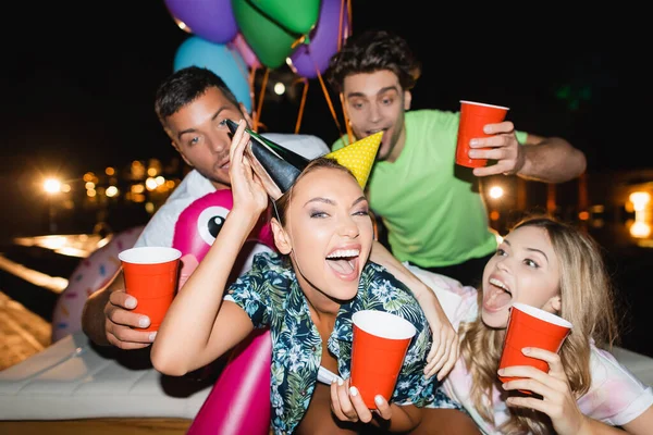 Selective focus of excited woman wearing party caps near friends with disposable cups and balloons outdoors at night — Stock Photo