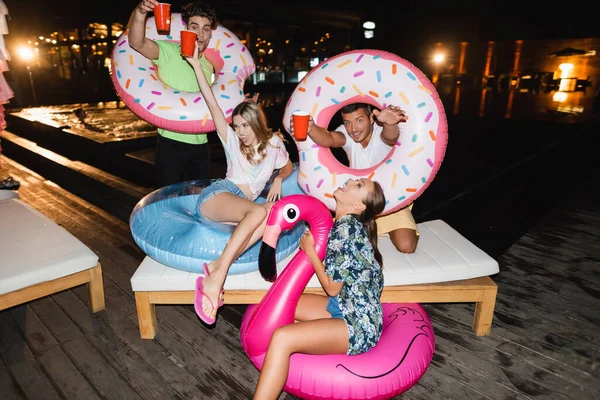 Amigos entusiasmados se divertindo com copos descartáveis e anéis de natação durante a festa perto da piscina à noite — Fotografia de Stock
